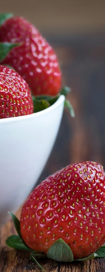 Close-up of fresh strawberries in a white bowl on a rustic wooden table, highlighting vibrant red color.