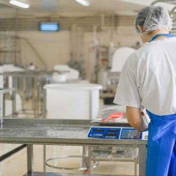A factory worker in uniform measures ingredients on a digital scale in an industrial setting.